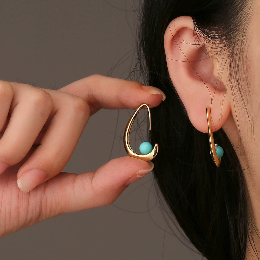 Close-up of person wearing and holding gold-plated earrings with turquoise stone, minimalistic jewelry design