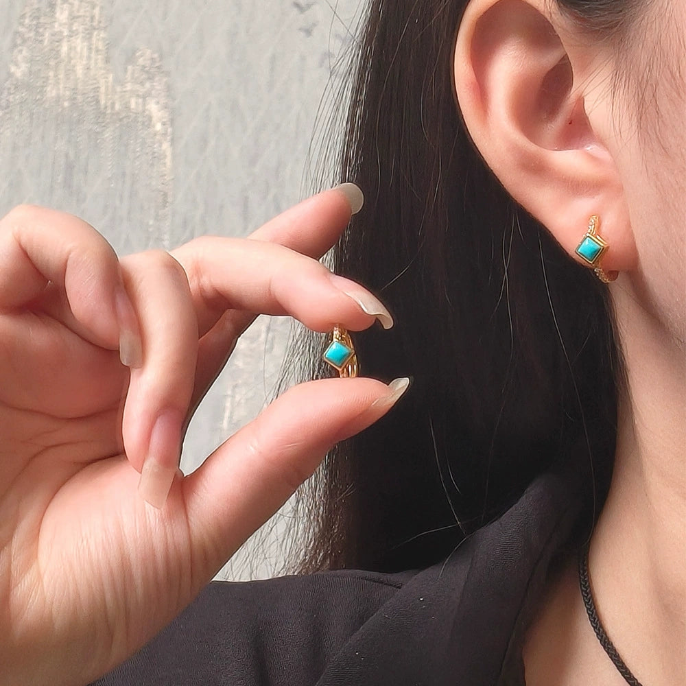 Close-up of a woman wearing and holding gold-plated earrings with square turquoise gemstones