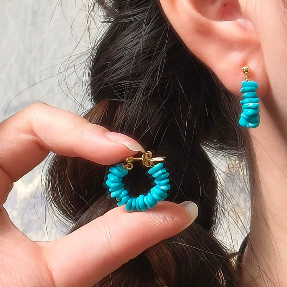 Close-up of a woman wearing turquoise stone and gold plated earrings, holding a matching turquoise hoop earring