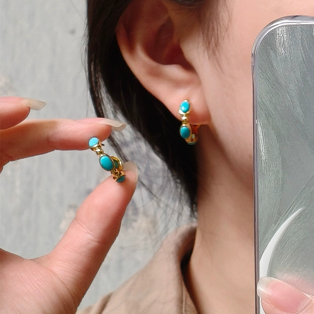 Close-up of woman wearing gold plated turquoise gemstone hoop earrings, holding matching earring near ear