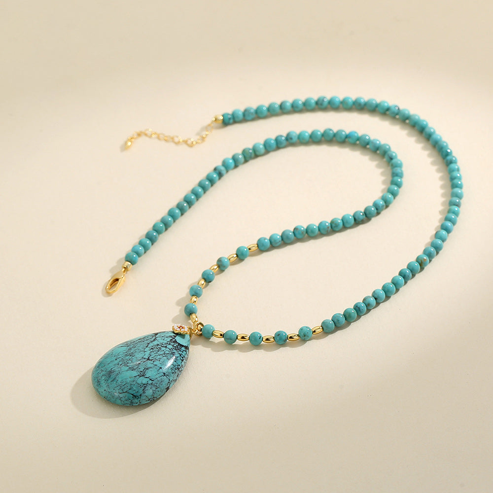 Turquoise bead necklace with gold accents and large teardrop gemstone pendant on beige background
