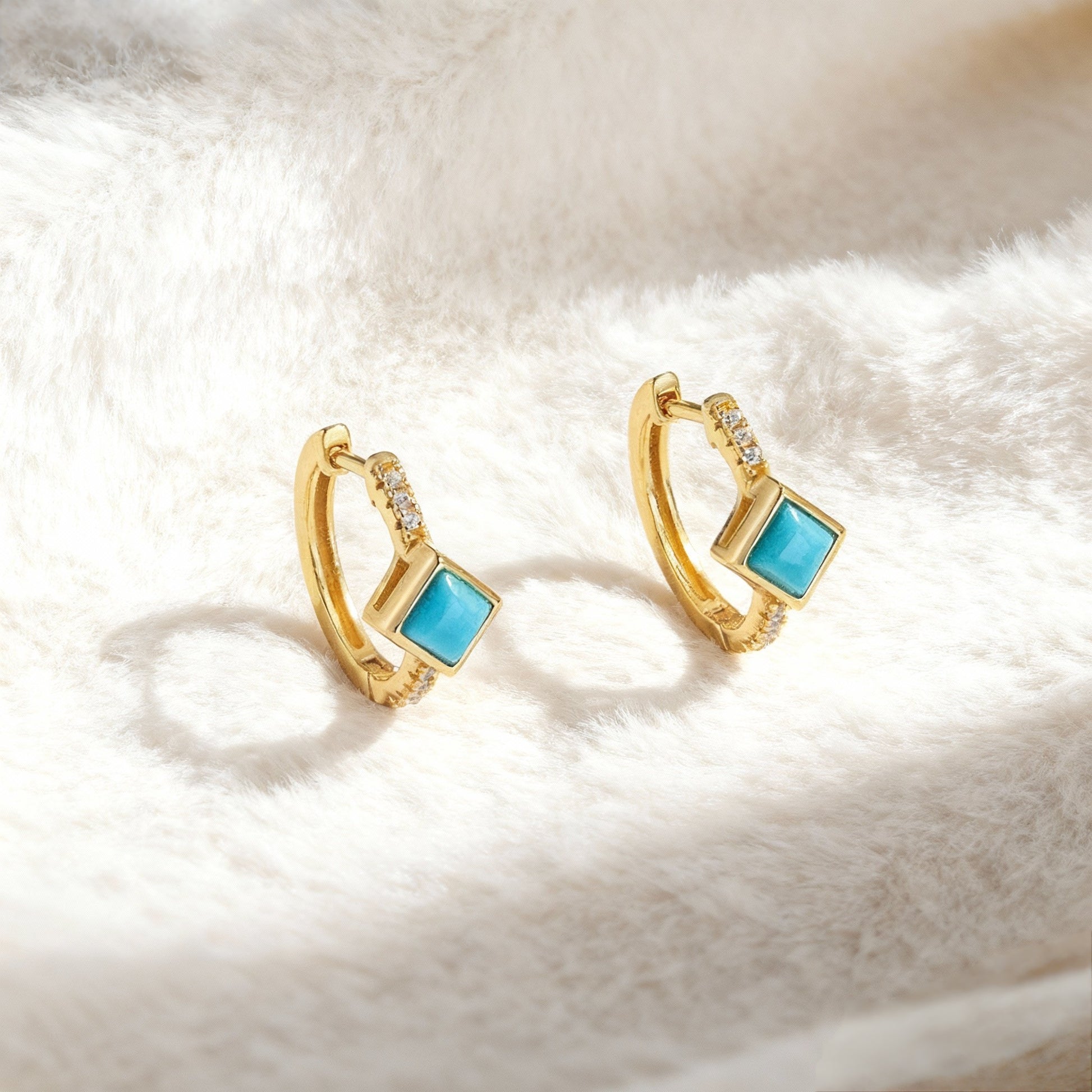 Gold plated hoop earrings with square turquoise gemstones and small embedded crystals on soft white fabric