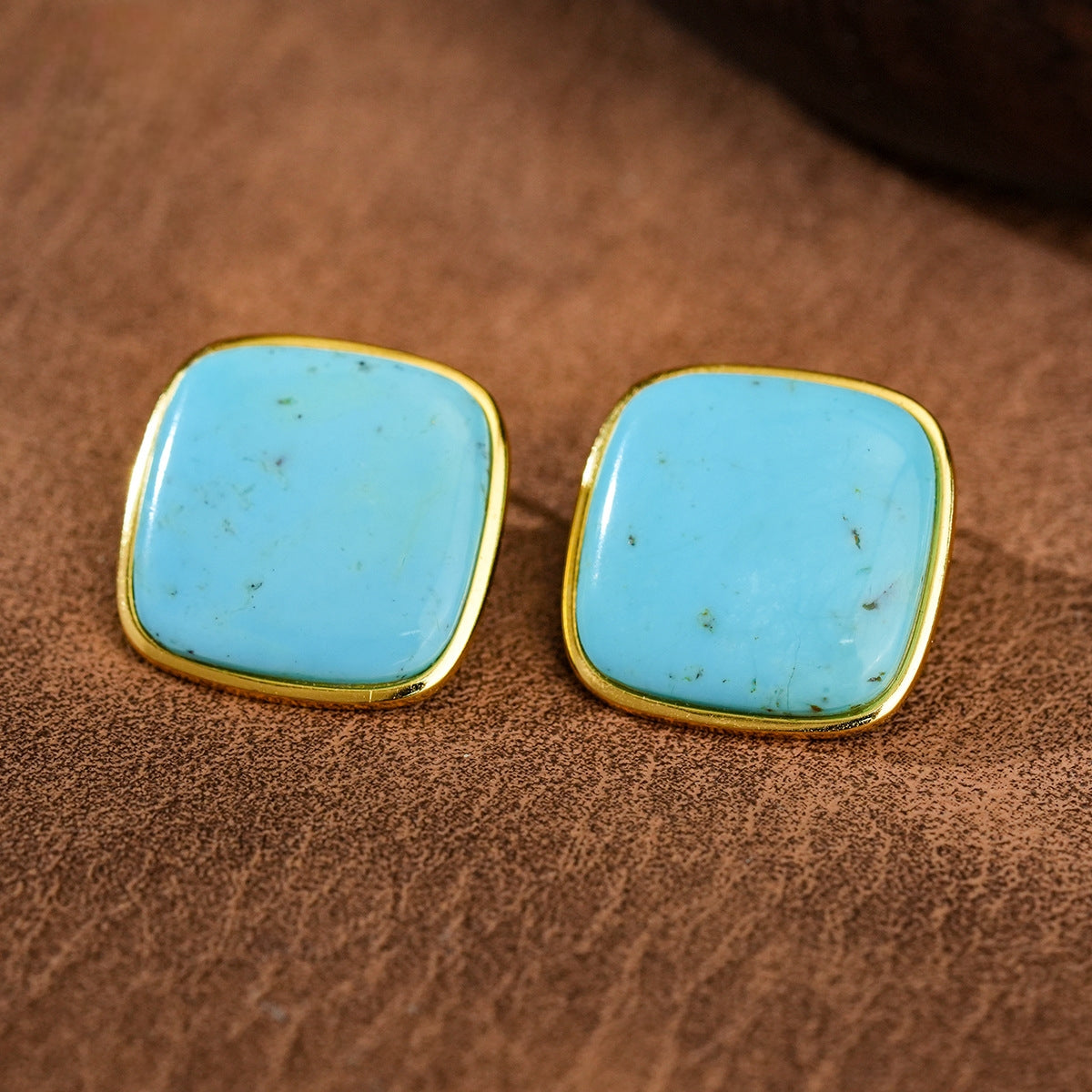 Square turquoise gemstone earrings with gold plated edges on brown textured surface